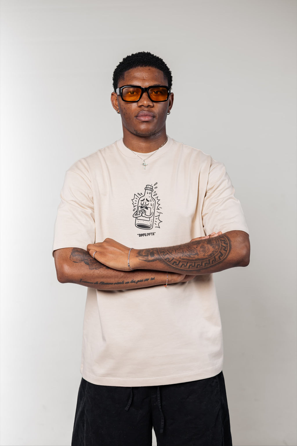 Man wearing a beige t-shirt with a graphic design, black pants, and sunglasses against a plain background